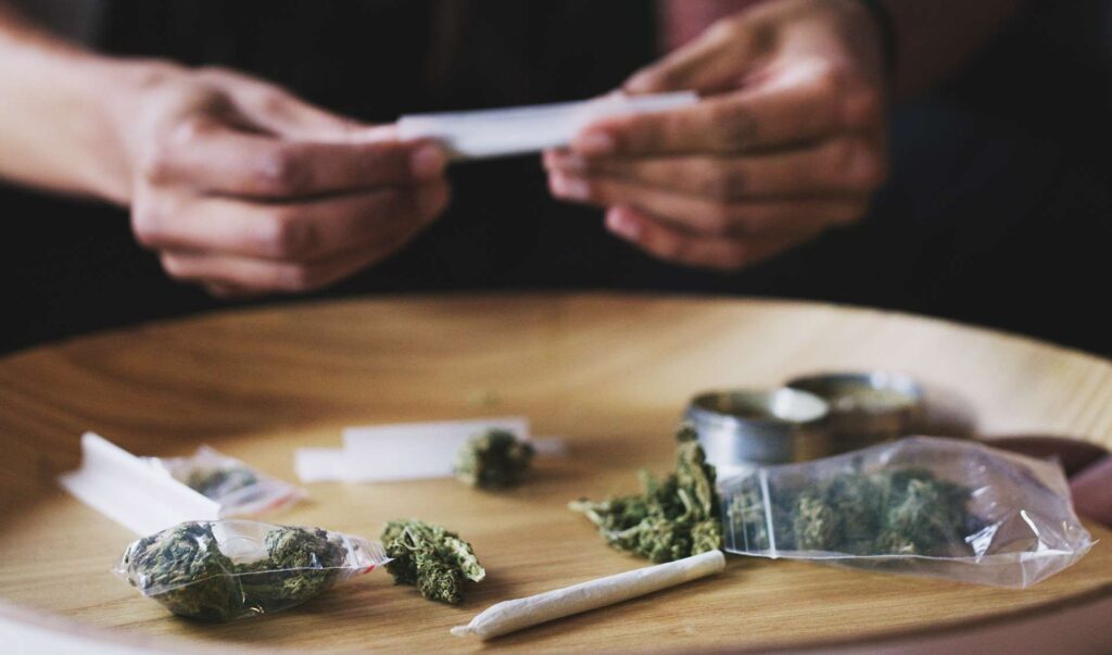 A person rolling a marijuana joint on a wooden table with cannabis buds, bags, and a grinder nearby.